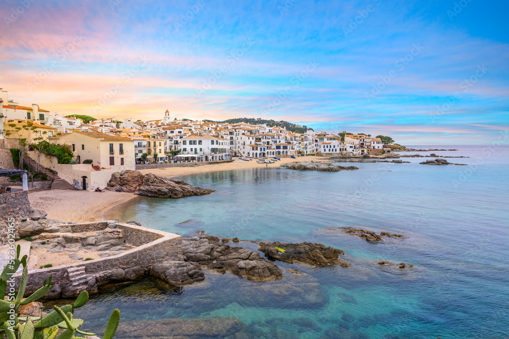 The whitewashed fishing village of Calella de Palafrugell along the sandy and rocky coastline of the Costa Brava under a colorful sky.