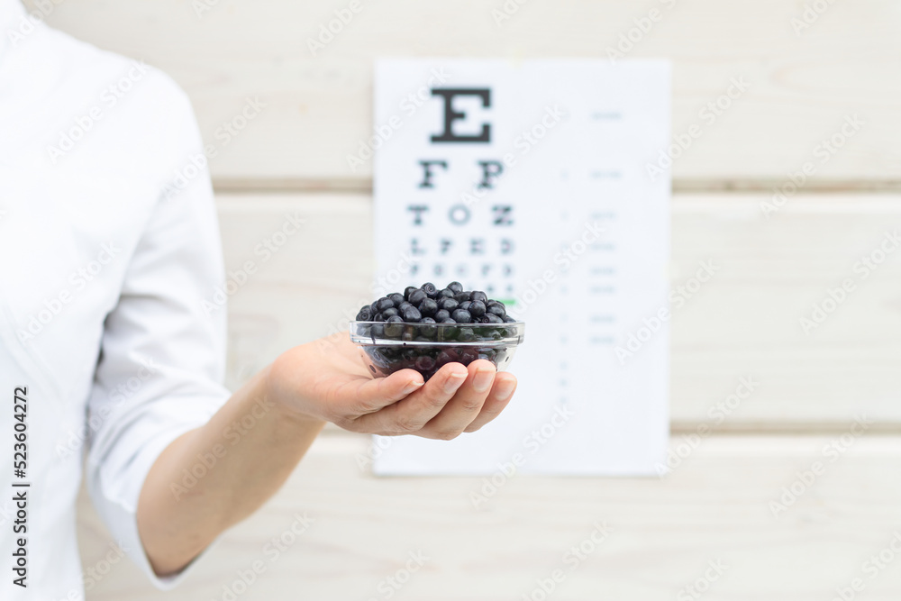 Obraz premium A bowl of blueberries in a doctor's female hand, Snellen's vision chart in the background. Natural vitamins for the eyes.