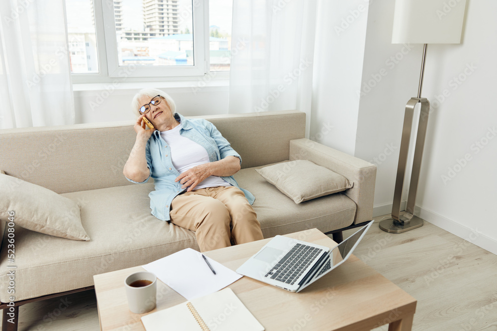 a happy, relaxed elderly woman with short white hair is sitting on a cozy sofa in an apartment and talking on a smartphone stylishly dressed in a light shirt and with glasses on her face a happy, relaxed elderly woman with short white hair is sitting on a cozy sofa in an apartment and talking on a smartphone stylishly dressed in a light shirt and with glasses on her face