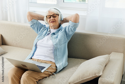 a relaxed elderly lady is watching a video with headphones, holding a laptop on her lap, leaning on the sofa and holding her hands behind her head © Tatiana