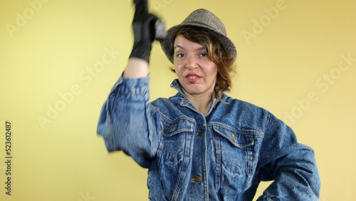 Forty-year-old woman in a denim jacket and hat shoots with a pistol on an isolated yellow background. Portrait of a bandit woman photo