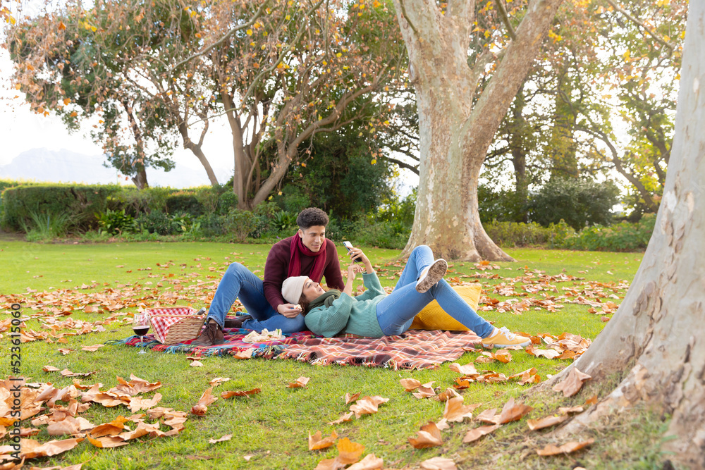 Fototapeta premium Happy biracial couple having picnic on a rug and using smartphone in autumn garden