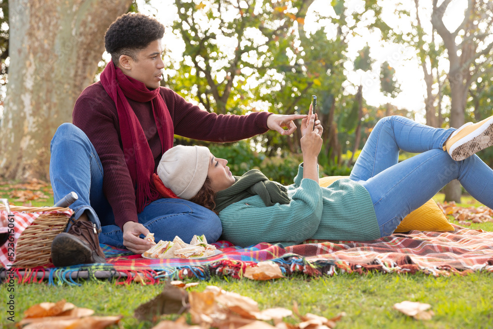 Fototapeta premium Happy biracial couple having picnic on a rug and using smartphone in autumn garden, smiling