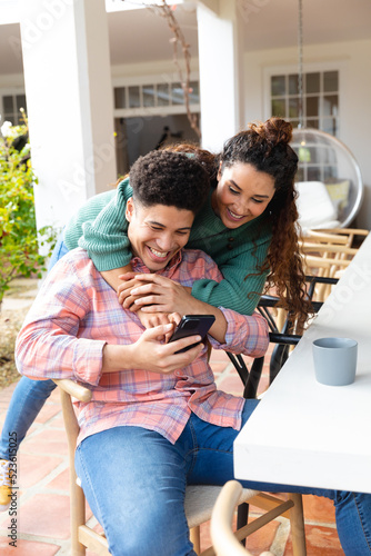 Happy biracial couple on garden terrace embracing outside house smiling and looking at smartphone