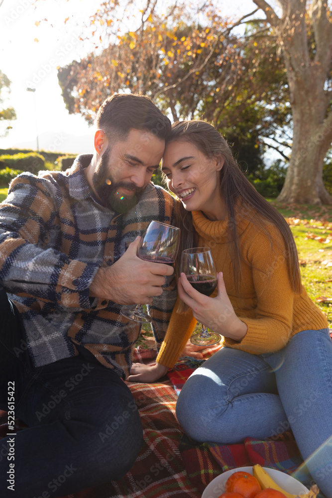 Fototapeta premium Romantic caucasian couple sitting on rug in sunny autumn garden drinking wine