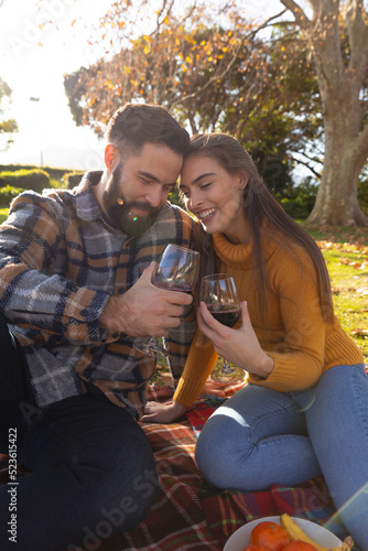 Romantic caucasian couple sitting on rug in sunny autumn garden drinking wine