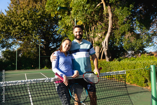 Portrait of happy caucasian couple playing tennis embracing by the net on sunny outdoor tennis court