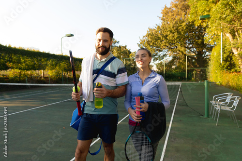 Smiling caucasian couple holding rackets walking on outdoor tennis court after playing tennis