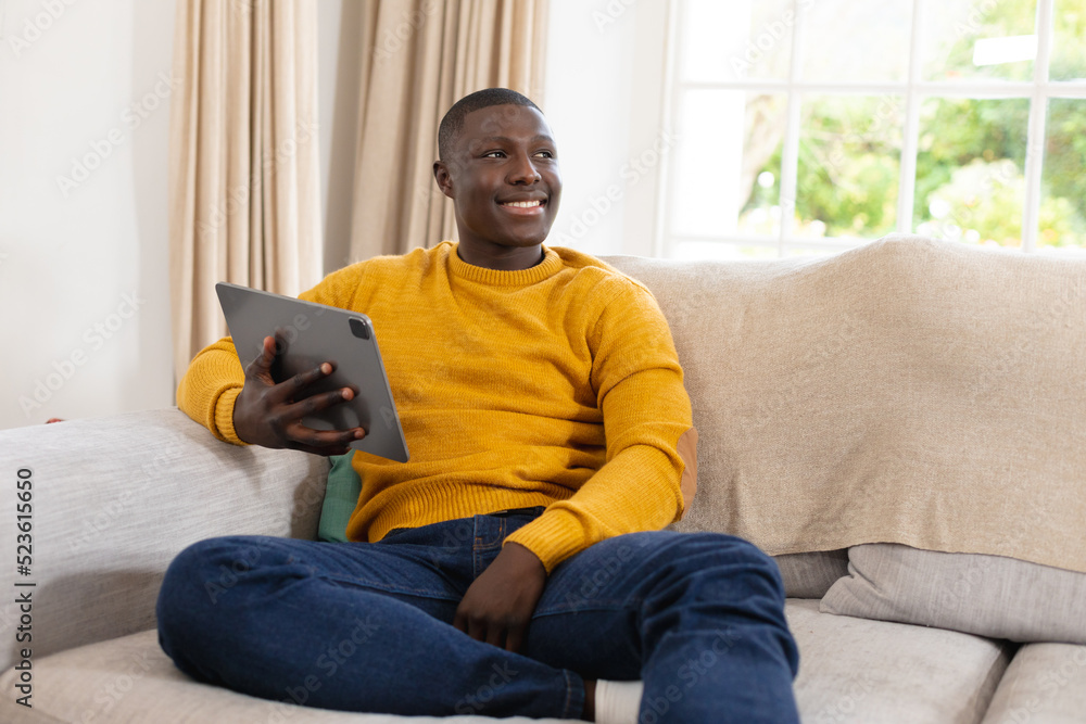 Smiling african american man sitting on couch at home holding tablet and looking away