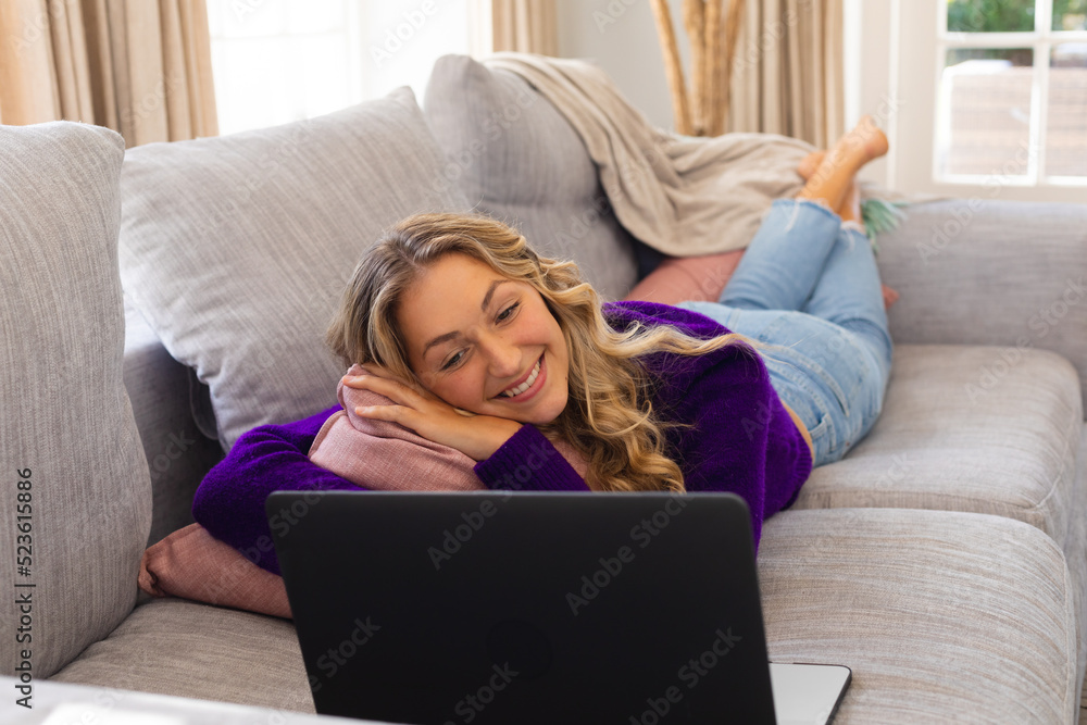 Smiling caucasian woman lying on couch looking at laptop in sunny living room Smiling caucasian woman lying on couch looking at laptop in sunny living room
