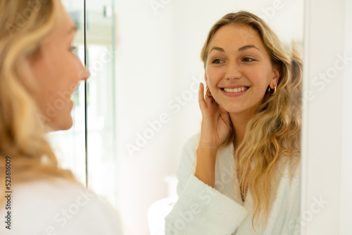 Happy caucasian woman wearing robe, looking at her reflection in mirror and smiling in bathroom