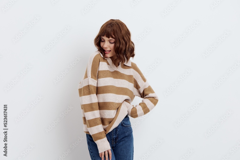 horizontal photo on a light background of a cute, pleasant woman in a stylish striped sweater correcting it on one side on the belt while standing in blue jeans