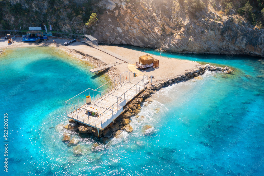 Aerial view of beautiful wooden pier, sea bay, sandy beach at sunset in summer. Top view of jetty, transparent blue water with waves, stones in lagoon, people. Tropical landscape. Vacation in Turkey