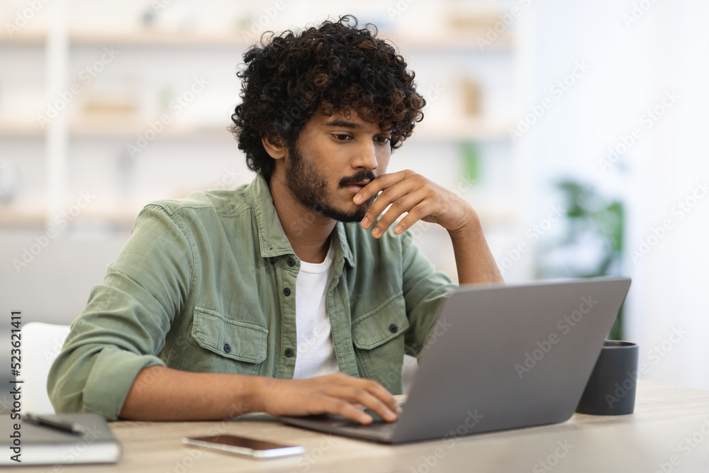 Pensive dark-skinned guy sitting at workdesk in front of laptop Pensive dark-skinned guy sitting at workdesk in front of laptop