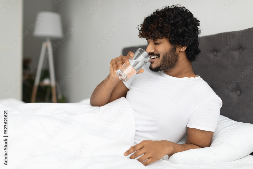 Happy young indian man holding glass of water in bed Happy young indian man holding glass of water in bed
