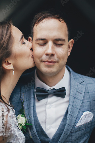 Stylish bride kissing happy groom in cheek on background of old church. Romantic moment. Provence wedding. Portrait of beautiful sensual wedding couple gently embracing in european city.