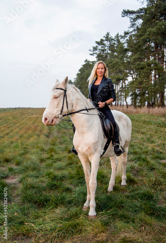 young beautiful blond smiling woman with long hair in black dress riding a white horse with blue eyes in autumn field 