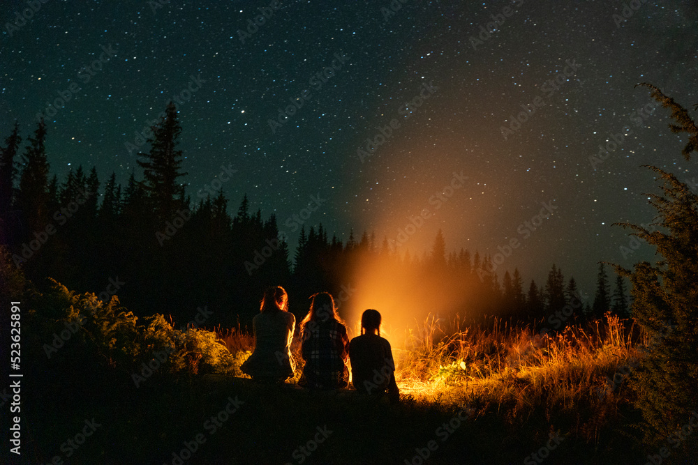 Lovely family sitting near the fireplace under the stars. Fire burning at night in a forest. Long exposure. Relaxing under the stars near campfire. Travel and adventure activity.