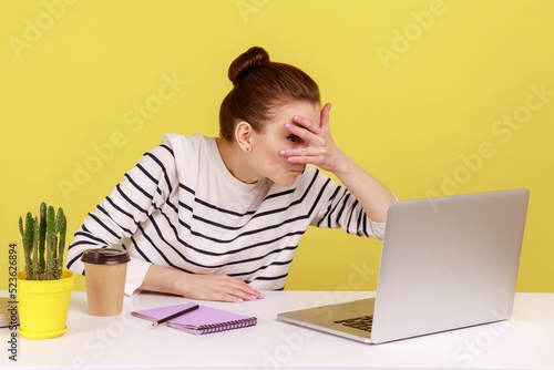 Curious woman manager sitting at workplace, spying, looking through fingers at laptop screen, peeking secret gossip in office. Indoor studio studio shot isolated on yellow background. photo