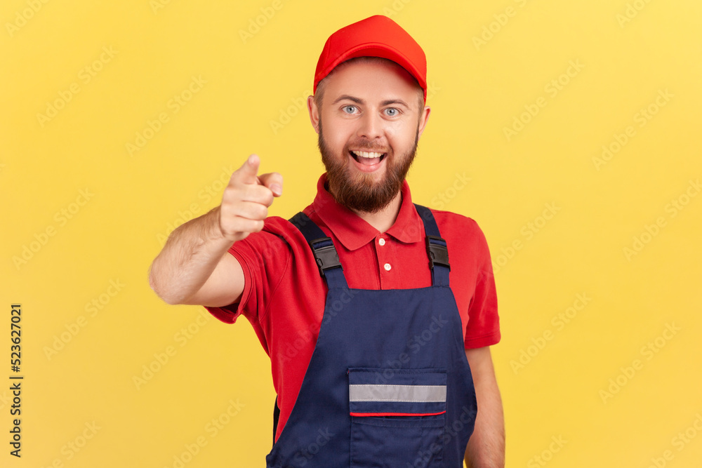 Portrait of smiling bearded satisfied man worker standing and pointing finger to camera, choosing you, wearing overalls and red cap. Indoor studio shot isolated on yellow background.