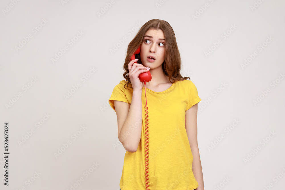 Portrait of amazed young curly haired female in yellow casual style T-shirt talking vintage phone, surprised by conversation, looking away. Indoor studio shot isolated on gray background. Portrait of amazed young curly haired female in yellow casual style T-shirt talking vintage phone, surprised by conversation, looking away. Indoor studio shot isolated on gray background.