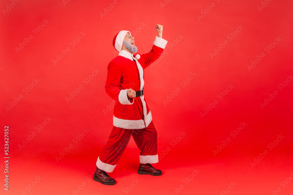 Full length portrait of elderly man with gray beard wearing santa claus costume screaming for joy with raised fists, celebrating holiday, champion. Indoor studio shot isolated on red background. Full length portrait of elderly man with gray beard wearing santa claus costume screaming for joy with raised fists, celebrating holiday, champion. Indoor studio shot isolated on red background.