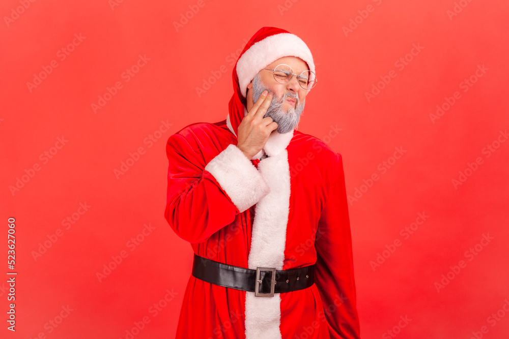 Portrait of sick elderly man with gray beard wearing santa claus costume standing and touching his cheek, suffering from terrible toothache. Indoor studio shot isolated on red background. Portrait of sick elderly man with gray beard wearing santa claus costume standing and touching his cheek, suffering from terrible toothache. Indoor studio shot isolated on red background.