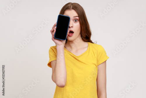 Portrait of excited shocked teenager girl wearing yellow T-shirt covering half face with cell phone and looking at camera with open mouth and big eyes. Indoor studio shot isolated on gray background.