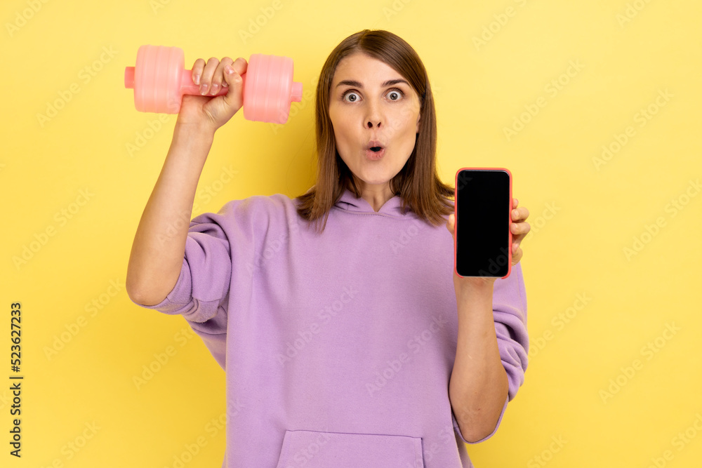 Shocked surprised woman holding blank screen smartphone and pink dumbbell, advertising new fitness application, wearing purple hoodie. Indoor studio shot isolated on yellow background. Shocked surprised woman holding blank screen smartphone and pink dumbbell, advertising new fitness application, wearing purple hoodie. Indoor studio shot isolated on yellow background.