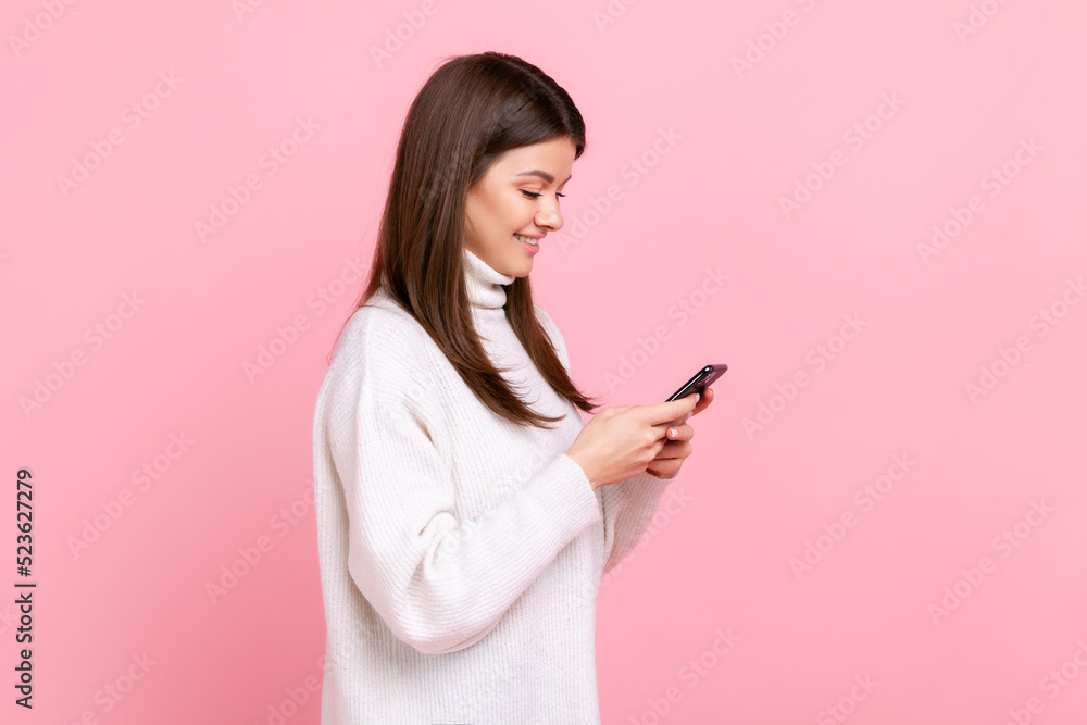 Side view of portrait optimistic girl using smartphone with happy expression, hearing pleasant news, wearing white casual style sweater. Indoor studio shot isolated on pink background. Side view of portrait optimistic girl using smartphone with happy expression, hearing pleasant news, wearing white casual style sweater. Indoor studio shot isolated on pink background.