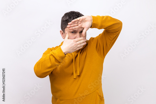 Frame of fingers. Portrait of man looking at camera through hand frame taking picture, focusing the eye, wearing urban style hoodie. Indoor studio shot isolated on white background.