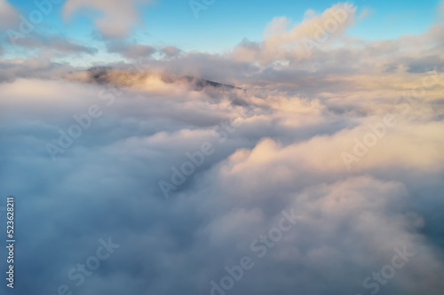 Mountains in clouds at sunrise in summer. Aerial view of mountain peak with green trees in fog. Top view from drone of mountain valley in low clouds