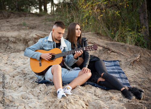 romantic couple sitting on the beach in autumn forest near lake and playing guitar © Tetatet