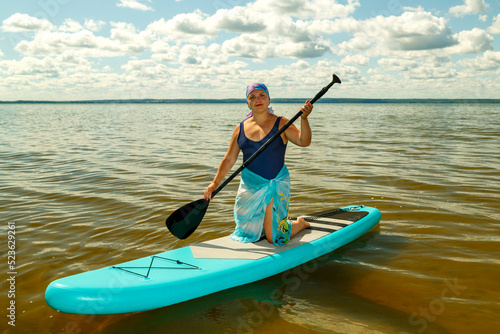A Jewish woman in a closed swimsuit, a pareo and a headdress on her knees on a SUP board with a paddle on the lake.