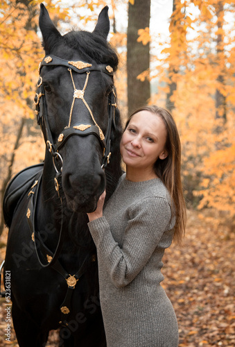 young beautiful smiling woman walking with black Friesian horse in autumn forest with yellow leaves