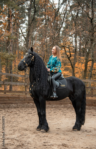 young beautiful smiling woman in ukrainian green embroidery riding black Friesian horse on a farm in autumn
