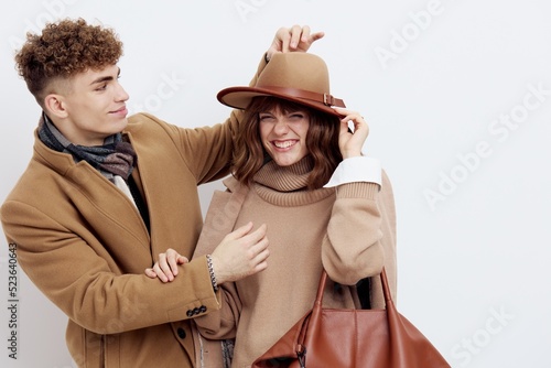 a cute couple is standing on a white background, wearing a coat. The man gently touches the top of the hat, the woman moves a little to the side, raising her hands to her head, holding the bag © Tatiana