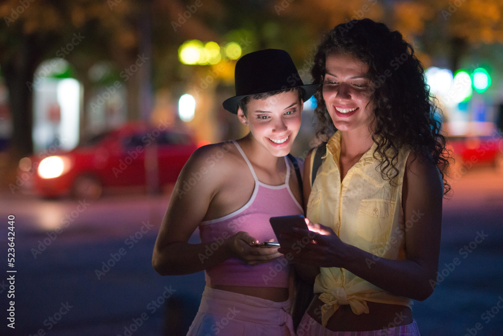 Young women using phone and talking while going out in town at summer night