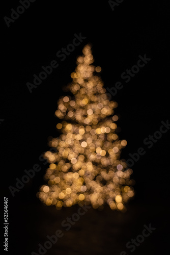 Silhouette of blurry Christmas tree illuminated and decorated with golden fairy lights in dark night creating beautiful bokeh effect with glowing circles or shiny dots, abstract image for holiday card