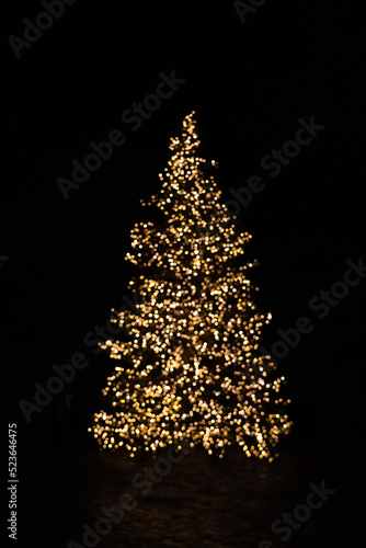Silhouette of blurry Christmas tree illuminated and decorated with golden fairy lights in dark night creating beautiful bokeh effect with glowing circles or shiny dots, abstract image for holiday card