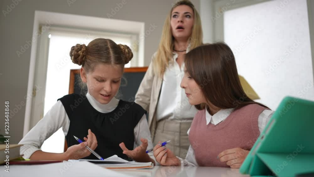 Hyperactive teen schoolgirls talking arguing sitting at desk in classroom with blurred teacher sighing standing at background. Caucasian girls quarreling in classroom in school with tired tutor Hyperactive teen schoolgirls talking arguing sitting at desk in classroom with blurred teacher sighing standing at background. Caucasian girls quarreling in classroom in school with tired tutor