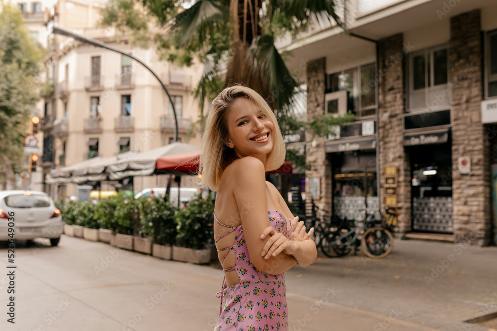Smiling caucasian lady blonde walks in sunny weather outdoors on city background with trees. Girl wears bright sundress. Sincere emotions lifestyle concept