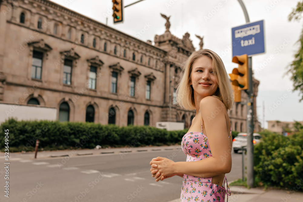 Modern pretty woman with short stylish hairstyle wearing summer dress is having fun in the city in warm sunny day. Attractive happy woman stands on background of old city building 