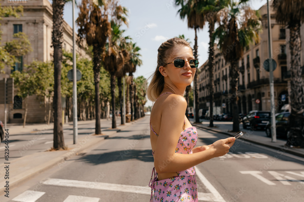 Pretty young european woman with smartphone in her hands outdoors. Blond with flowing hair walking on city with exotic plants on background. People sincere emotions lifestyle concept.