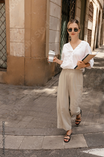 Obraz na płótnie Full-lenght portrait of smiling european pretty girl in sunglasses walking with coffee and magazines in the city. Blond wearing white shirt and beige pants walking on heels w salonie