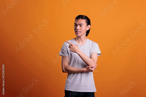 Young smiling confident asian man pointing to side with forefinger, advertising product. Cheerful teenager standing, showing promotion, studio medium shot on orange background