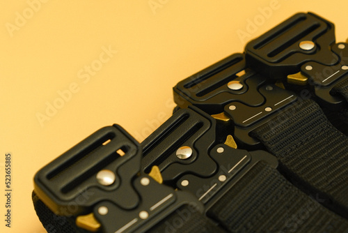 A side view of many black canvas belts with a black fast open metal male buckle and a black belt loop lays on the orange background. Utility. Hiking. Material. Outfit. Safety. Soldier. Steel. Weapon