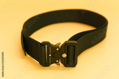 A black canvas belt with a black metal male buckle and a black belt loop lays on the orange background. Military. Buckle. Clothing. Equipment. Canvas. Protection. Tool. Strap. Metallic. War. Wear photo