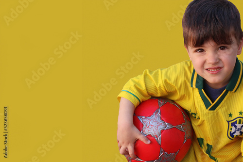 Menino com a camisa da seleção brasileira de futebol e fundo amarelo, segura em seus braços uma bola de futebol. photo