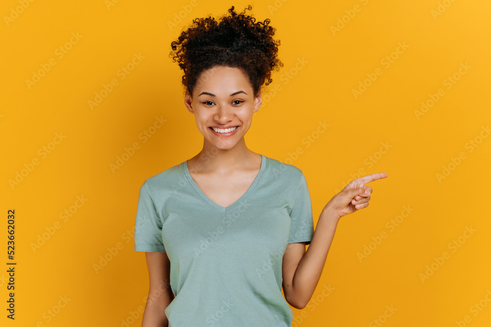 Lovely curly haired young african american woman in a basic t-shirt, with positive emotional facial expression, stand on isolated orange background, points finger to the side, looks at camera, smiles Lovely curly haired young african american woman in a basic t-shirt, with positive emotional facial expression, stand on isolated orange background, points finger to the side, looks at camera, smiles